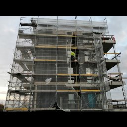Zakład ogolnobudowlany Rzepka Tomasz Stanisław - Budynek wielorodzinny w trakcie prac elewacyjnych, osłonięty siatką na rusztowaniach, z widocznym pracownikiem w żółtej kamizelce na wysokości.
