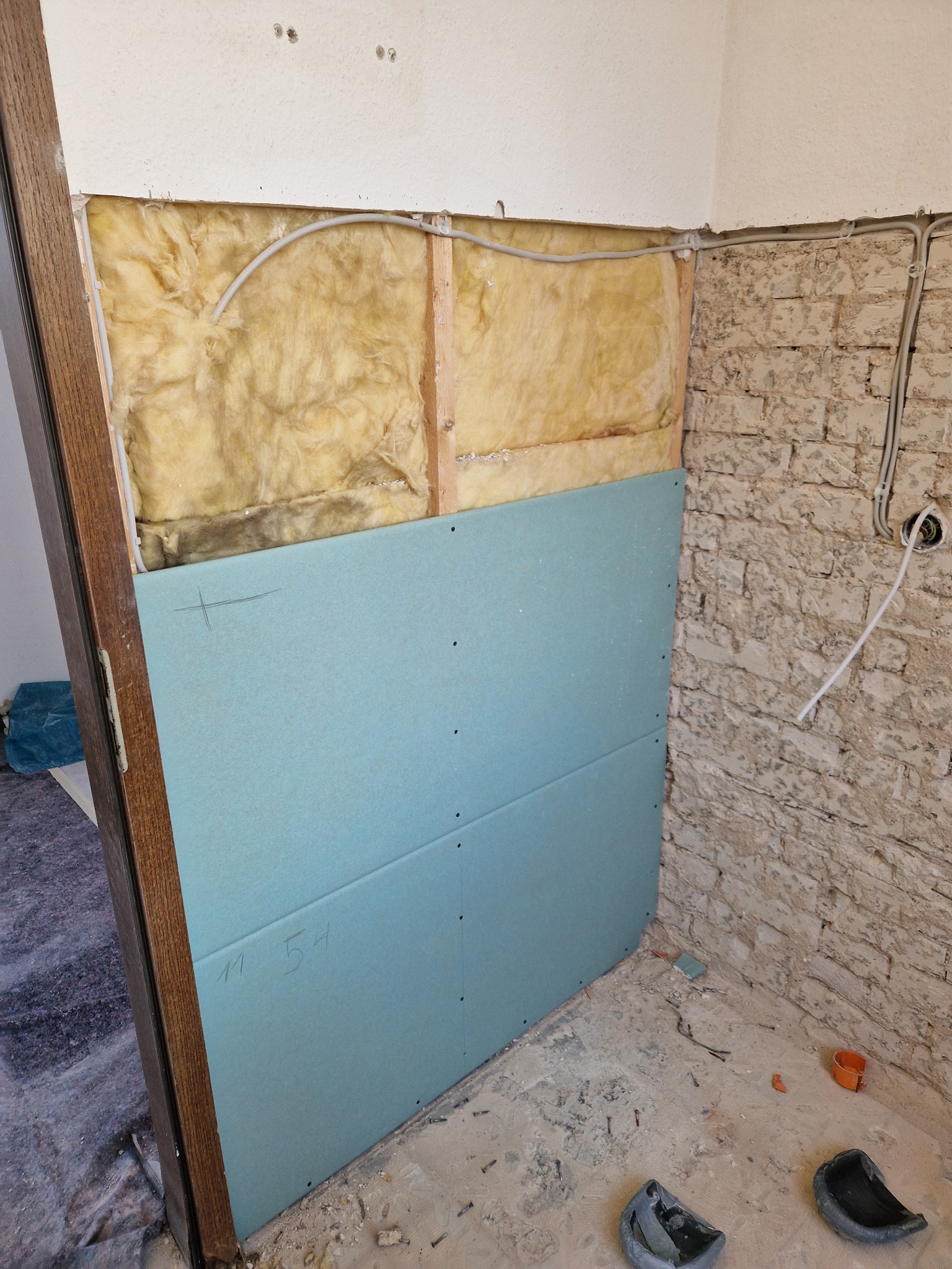 Wnętrze w trakcie prac: izolacja termiczna wełną mineralną, częściowo zakryta płytami gipsowo-kartonowymi z naniesionymi oznaczeniami, surowa ściana z cegły, widoczne przewody elektryczne...