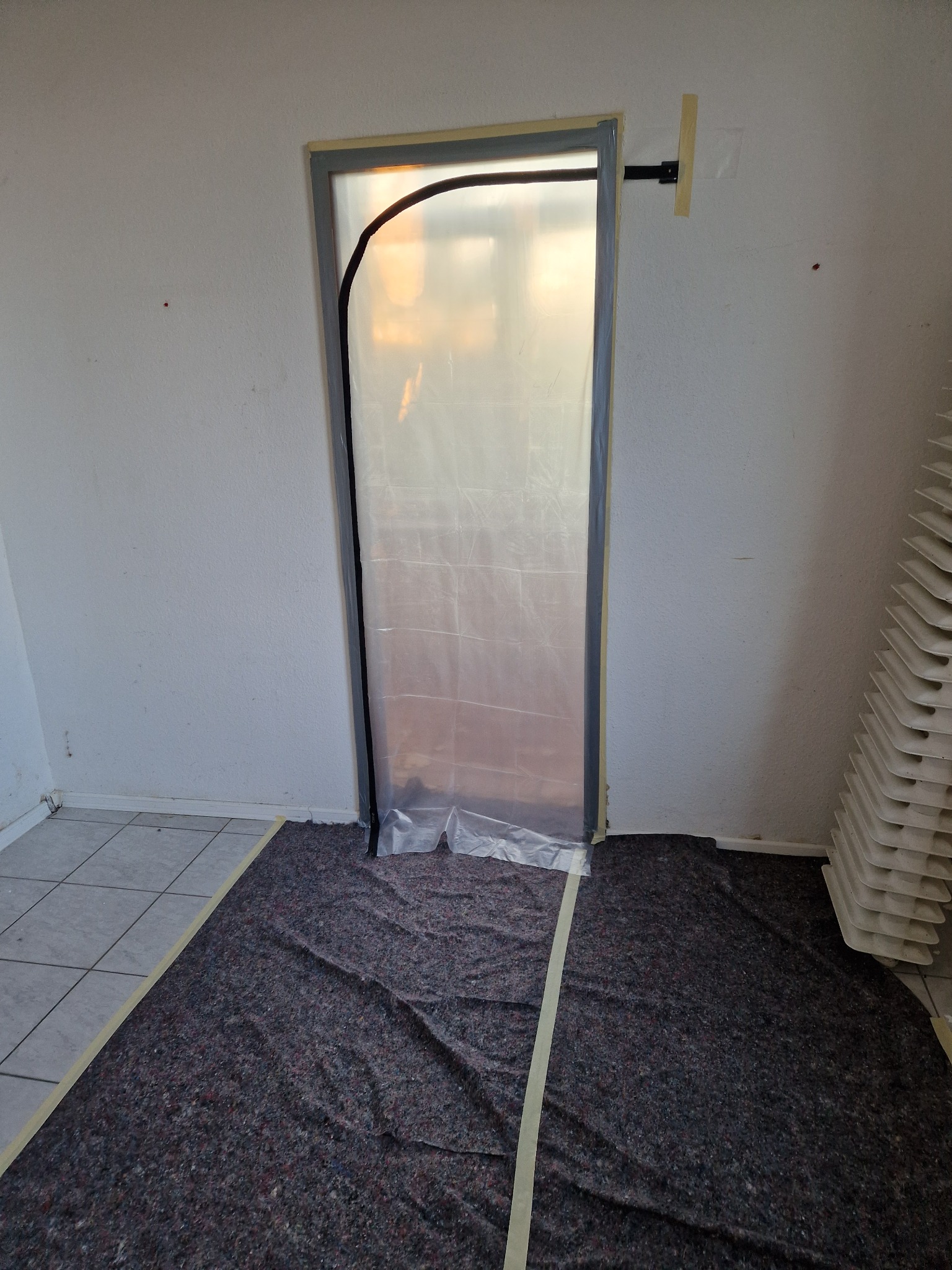 Przygotowanie do prac malarskich: zabezpieczone folią drzwi z czarną ramą i podłoga przykryta szarą matą, zabezpieczone taśmą malarską, widoczny grzejnik żeberkowy.