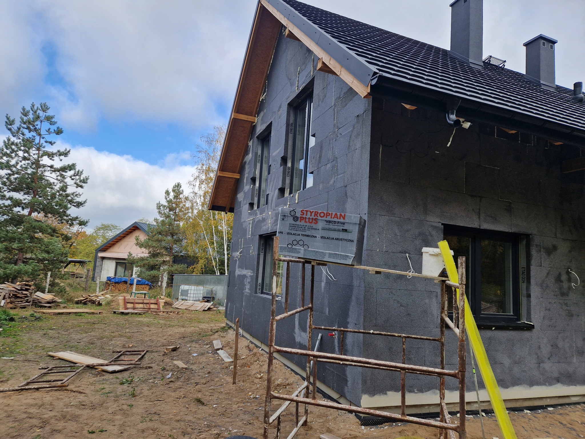 Dom w trakcie ocieplania elewacji, widoczny czarny styropian, rusztowanie i materiały budowlane na posesji.