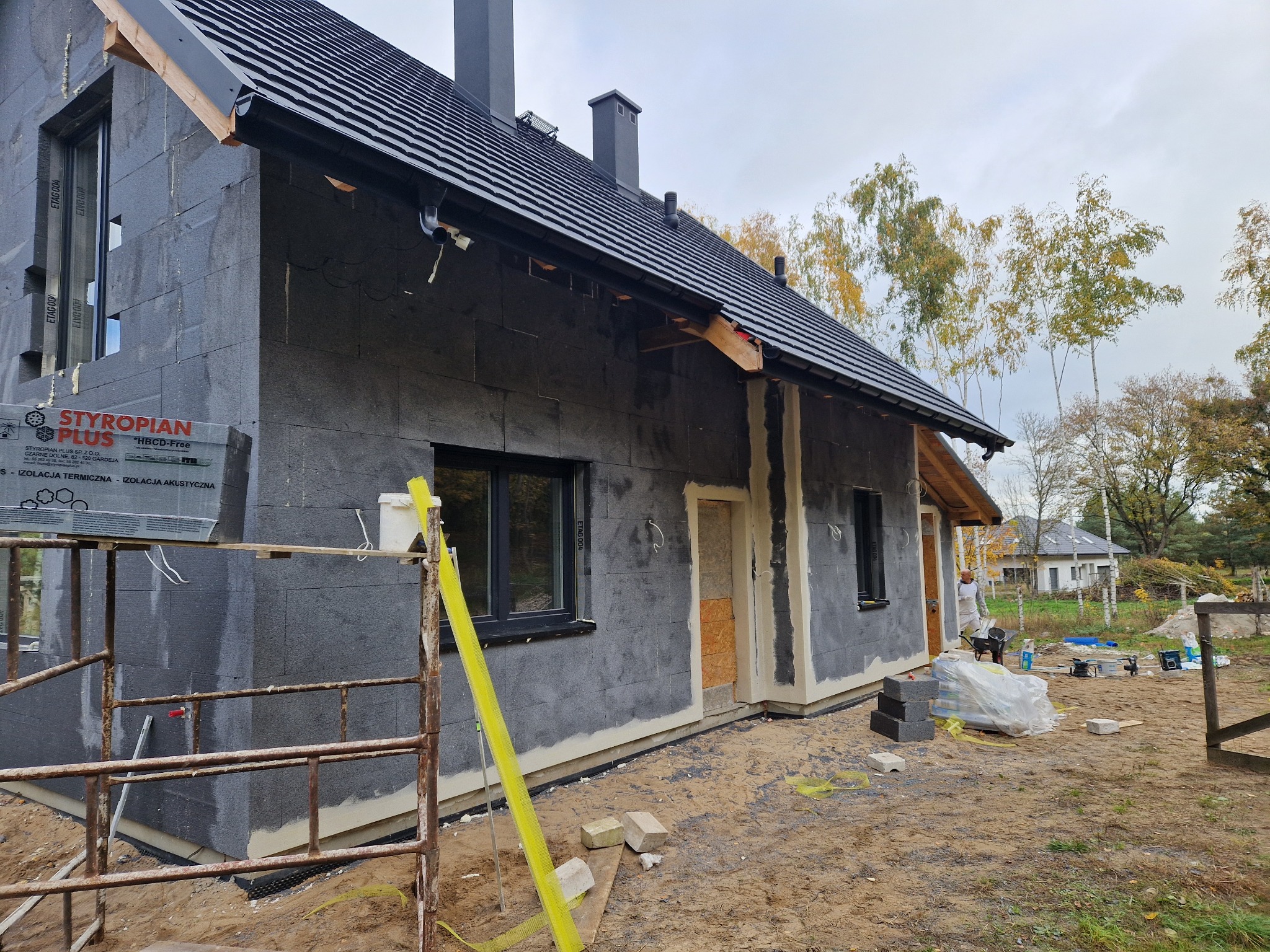 Dom w trakcie ocieplania elewacji styropianem grafitowym, widoczne rusztowania, paczki styropianu Styropian Plus, przygotowane wejście do budynku, teren budowy z piaskiem i częściowo ułożoną kostką.