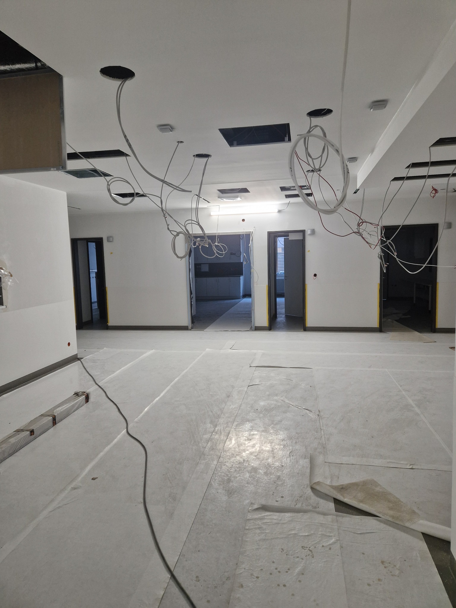 Wnętrze w trakcie prac wykończeniowych: zabezpieczona podłoga folią, widoczne kable zwisające z sufitu, przygotowane otwory na oświetlenie, trzy otwarte przejścia do kolejnych pomieszczeń.