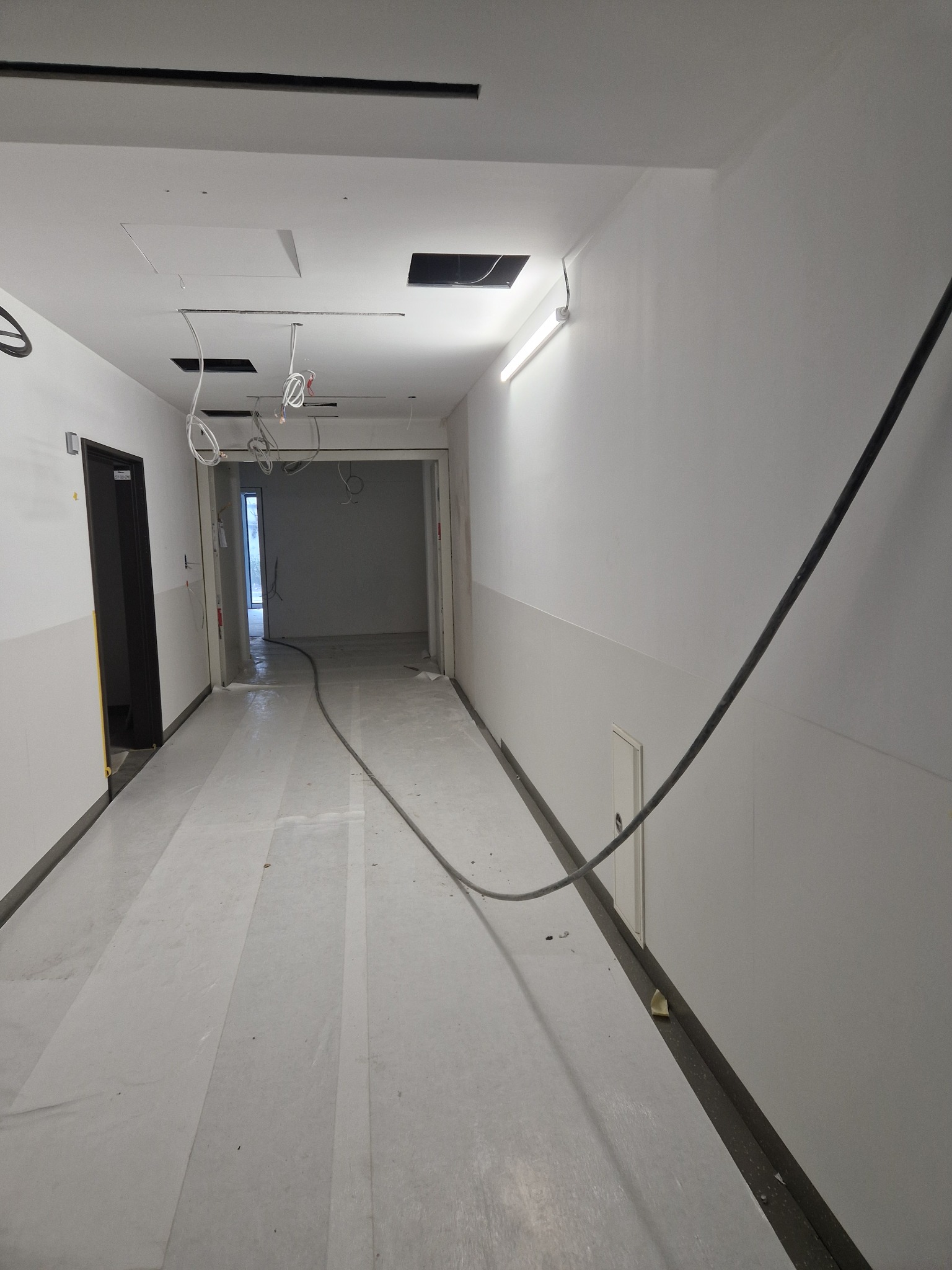 Korytarz w trakcie prac wykończeniowych, zabezpieczona podłoga folią, widoczne kable zwisające z sufitu i lampa fluorescencyjna na ścianie.