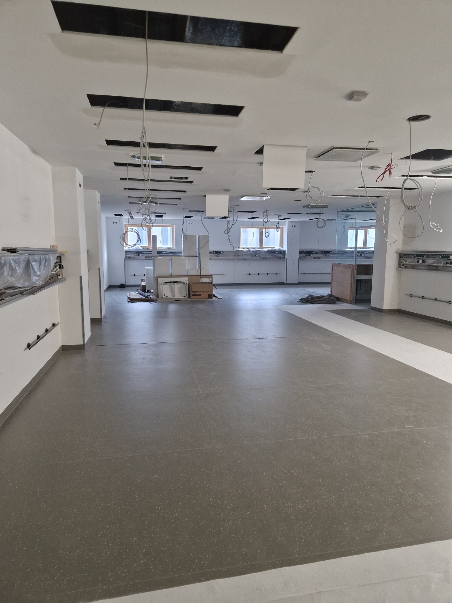 Wnętrze w trakcie remontu: szara podłoga, białe ściany, odsłonięty sufit z instalacjami i oknami w tle.