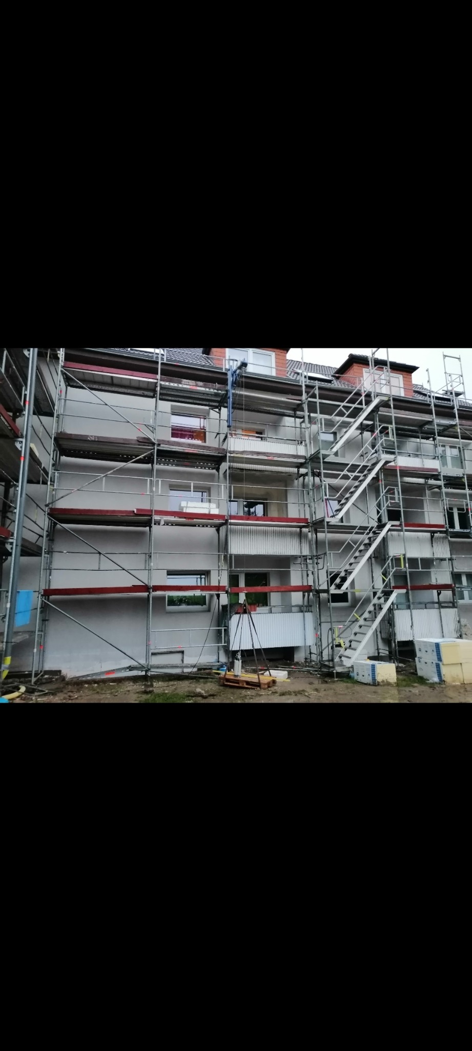 Wielopiętrowy budynek mieszkalny w trakcie ocieplania, z rusztowaniami i widocznymi płytami styropianowymi na palecie.