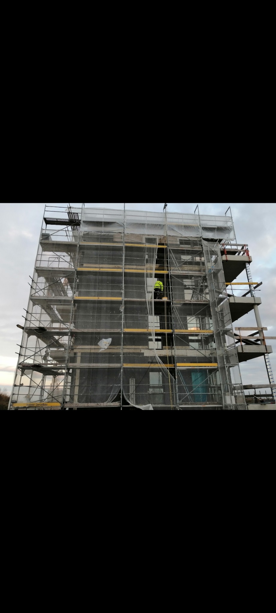 Budynek wielorodzinny w trakcie prac elewacyjnych, osłonięty siatką na rusztowaniach, z widocznym pracownikiem w żółtej kamizelce na wysokości.