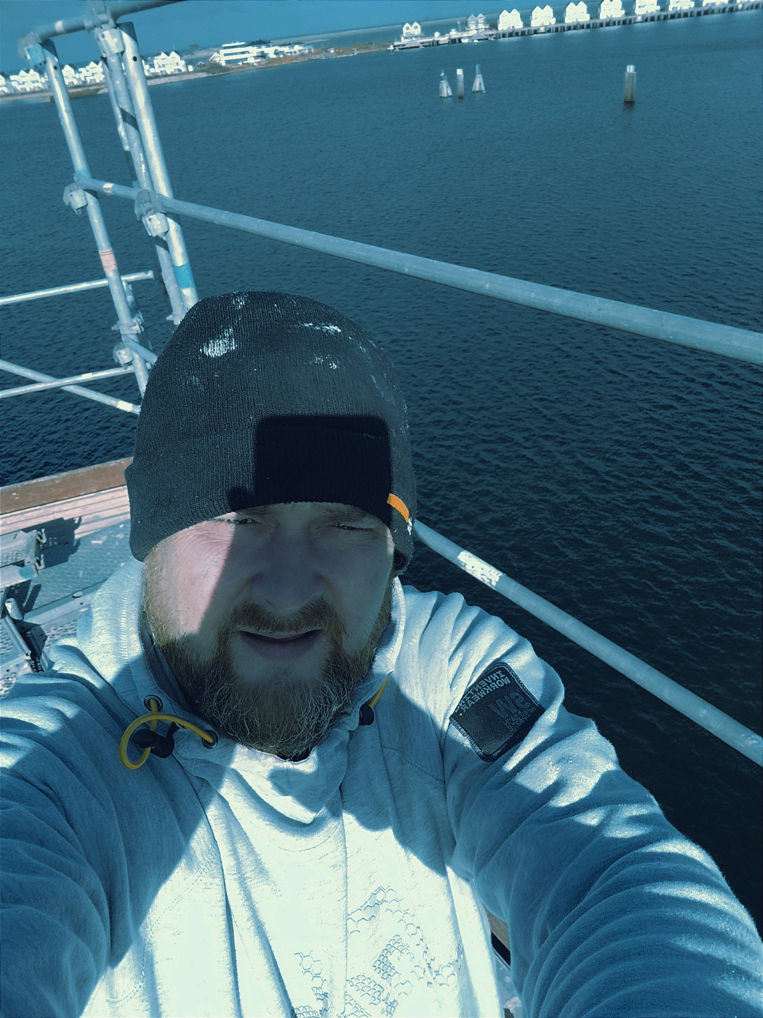 Mężczyzna w czapce i bluzie z kapturem robi selfie na rusztowaniu z widokiem na morze i zabudowania na brzegu.