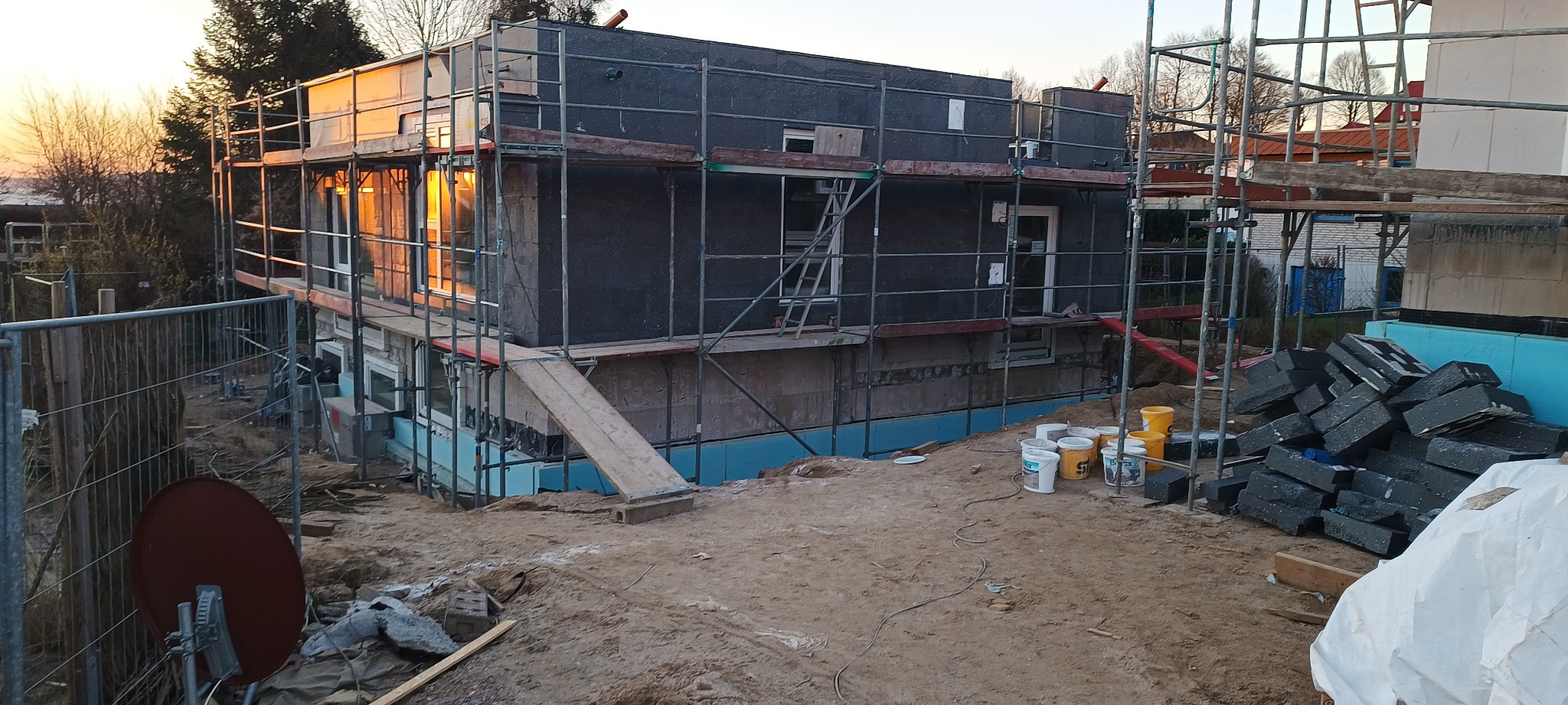 Budynek w trakcie ocieplania elewacji, widoczne rusztowania, styropianowe płyty ociepleniowe i wiaderka z farbą na placu budowy.