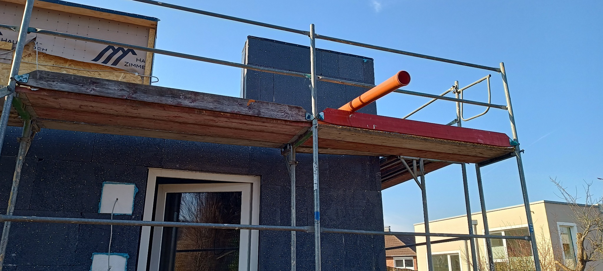 Elewacja budynku w trakcie ocieplania, widoczne rusztowanie, płyty izolacyjne z czarnym granulatem, okno z odbiciem drzew, pomarańczowa rura wentylacyjna wystająca z ocieplenia