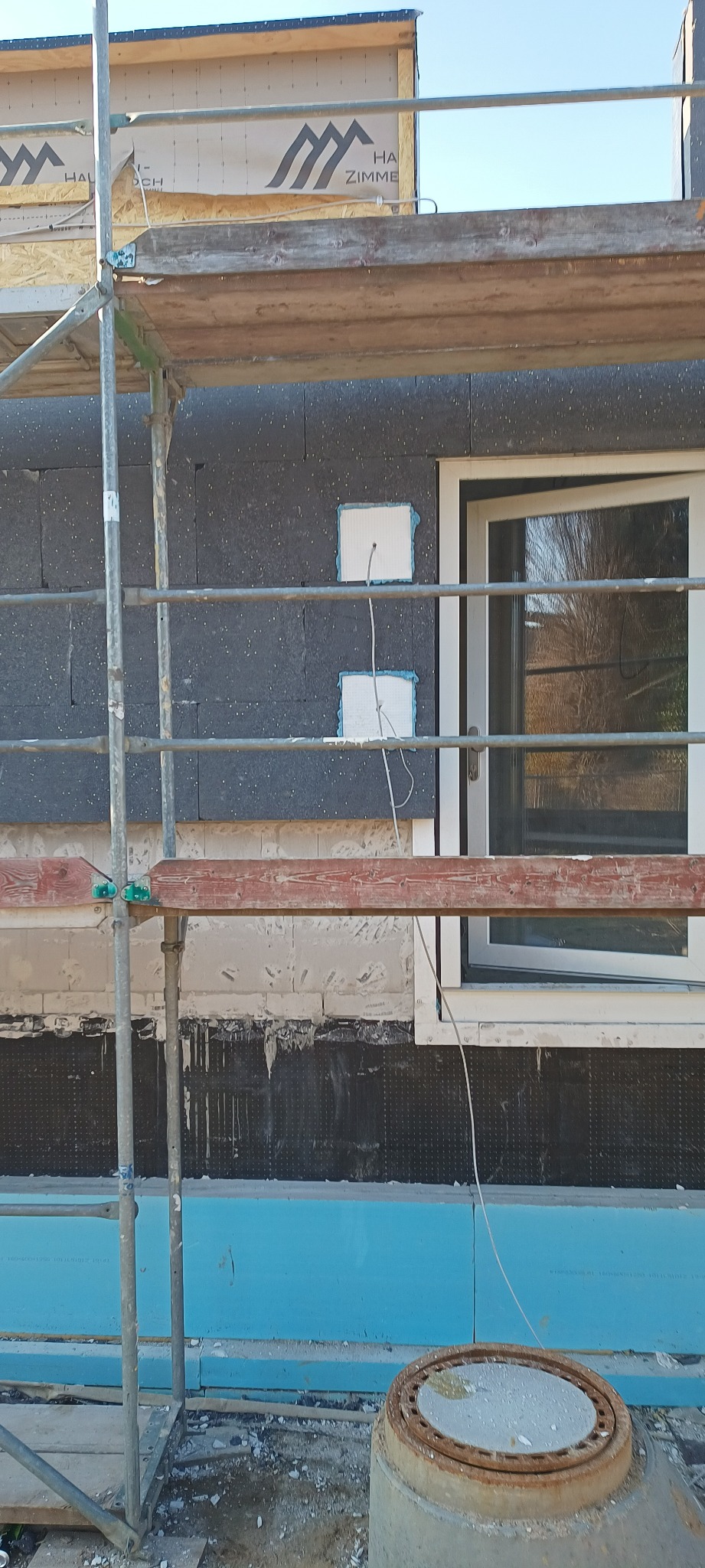 Fragment elewacji budynku w trakcie ocieplania, widoczne rusztowanie, warstwa izolacyjna z wyciętymi otworami na przewody, białe okno oraz betonowa studzienka kanalizacyjna w pobliżu ściany.