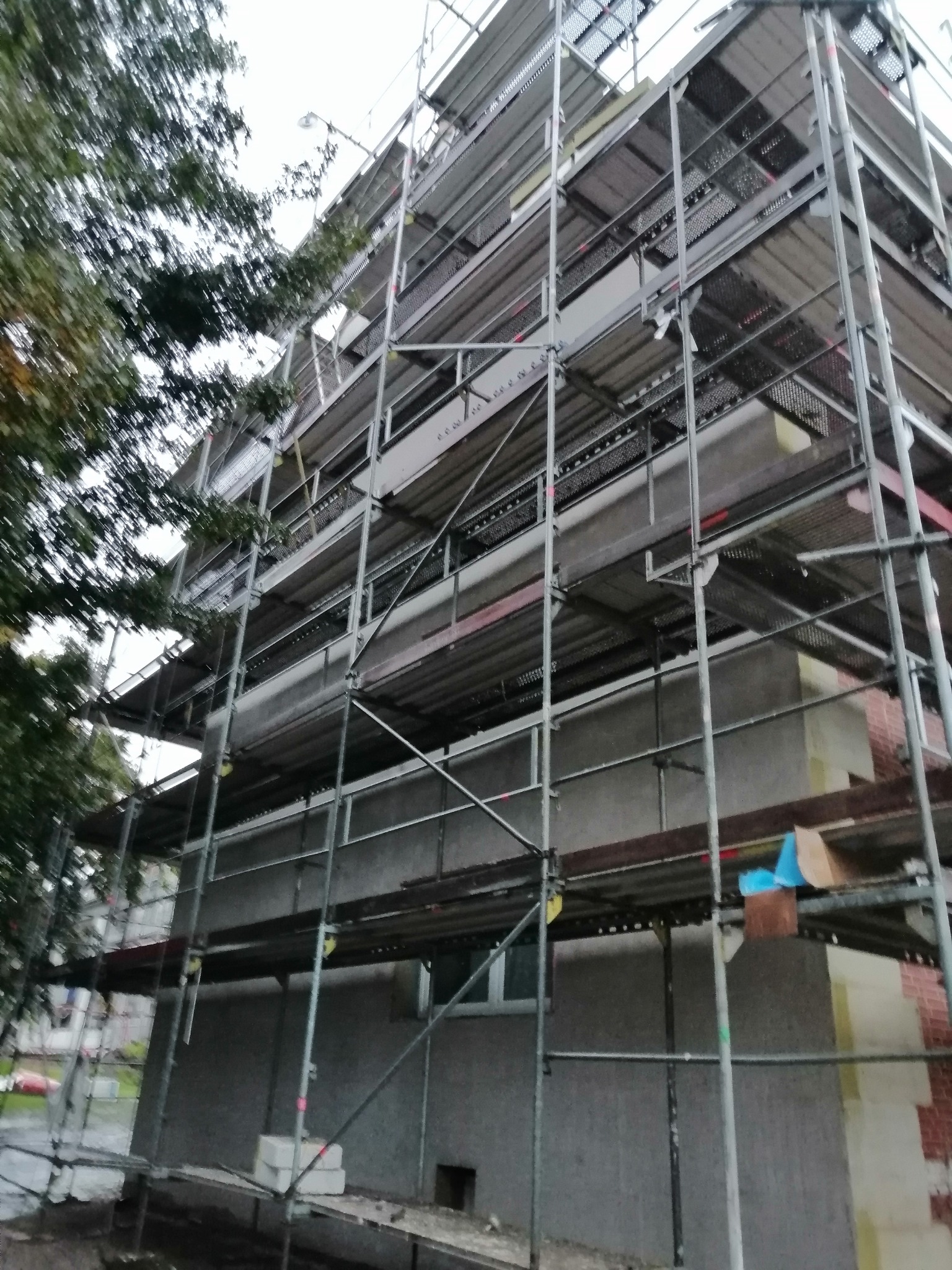 Budynek w trakcie ocieplania, widoczne rusztowania, szara elewacja i fragment ocieplenia styropianem, ujęcie z dołu.