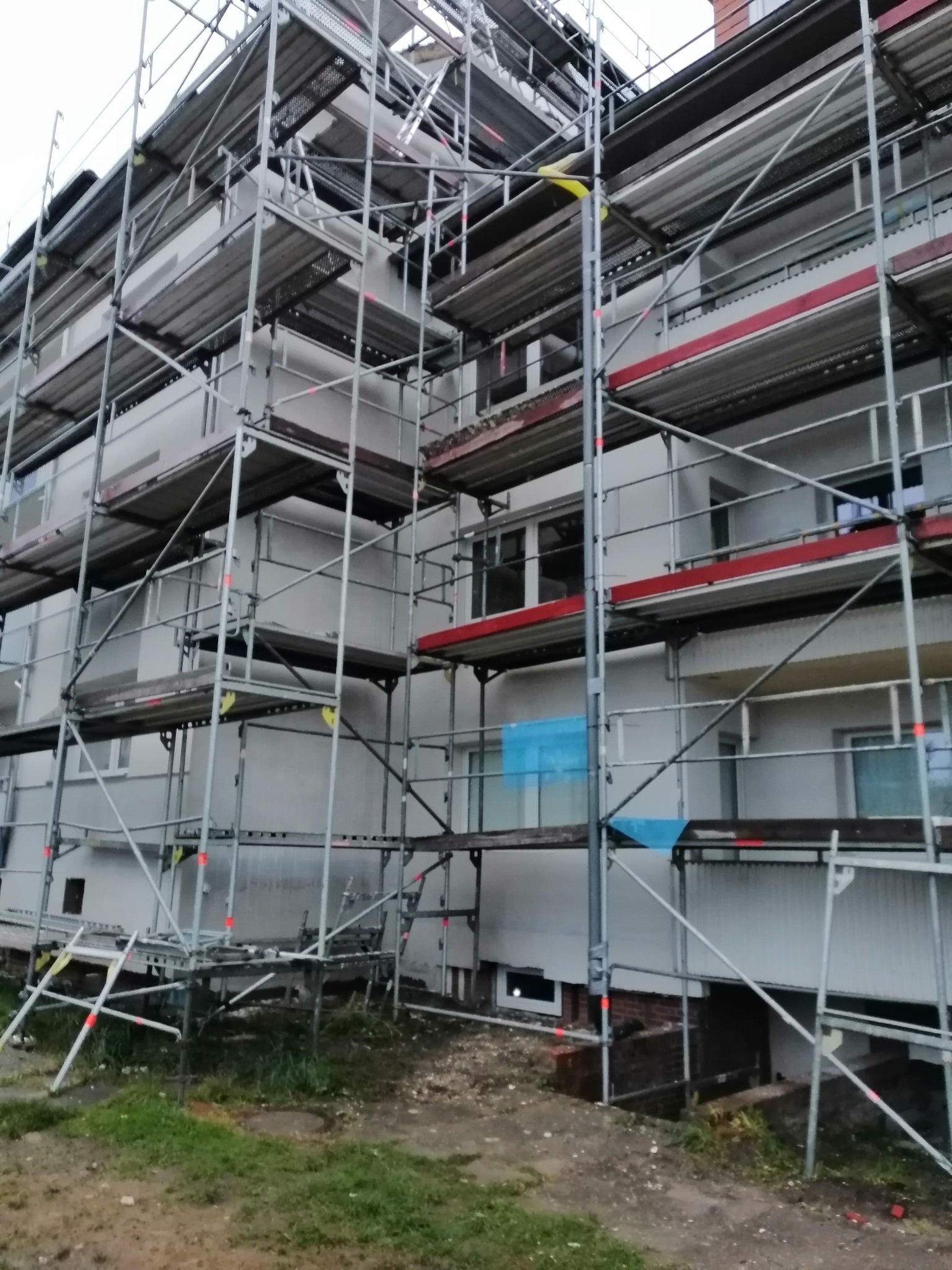 Wielopiętrowy budynek w trakcie ocieplania elewacji, widoczne rusztowania i fragmenty ocieplonej ściany z szarym tynkiem.