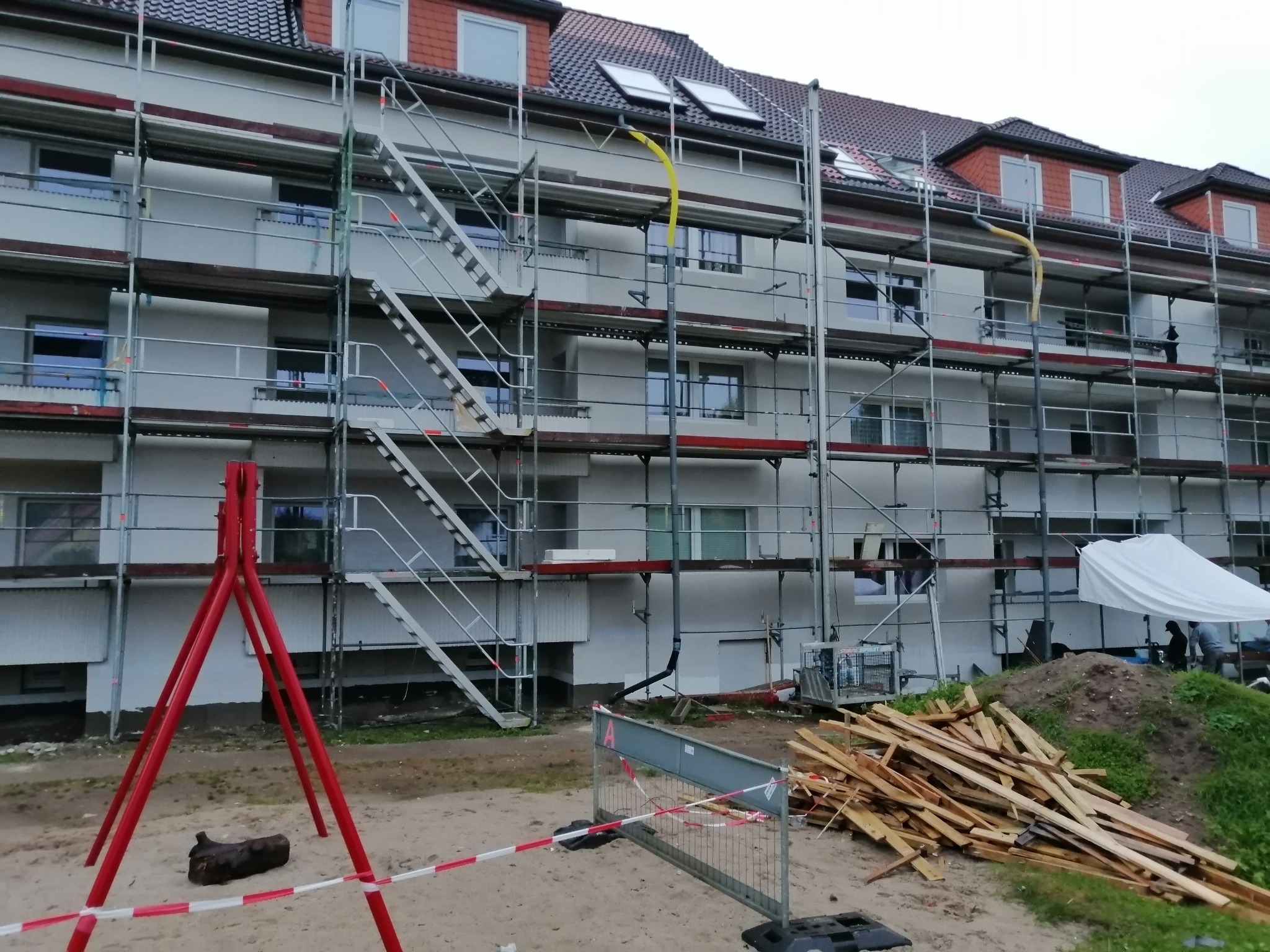 Wielopiętrowy budynek mieszkalny w trakcie prac elewacyjnych, otoczony rusztowaniami i zabezpieczeniami, z widocznym stosem drewna i elementami konstrukcyjnymi na pierwszym planie.