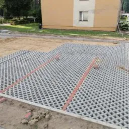 Układanie ażurowej kostki brukowej na przygotowanym podłożu piaskowym, widoczne czerwone listwy oddzielające sekcje, budynek w tle.