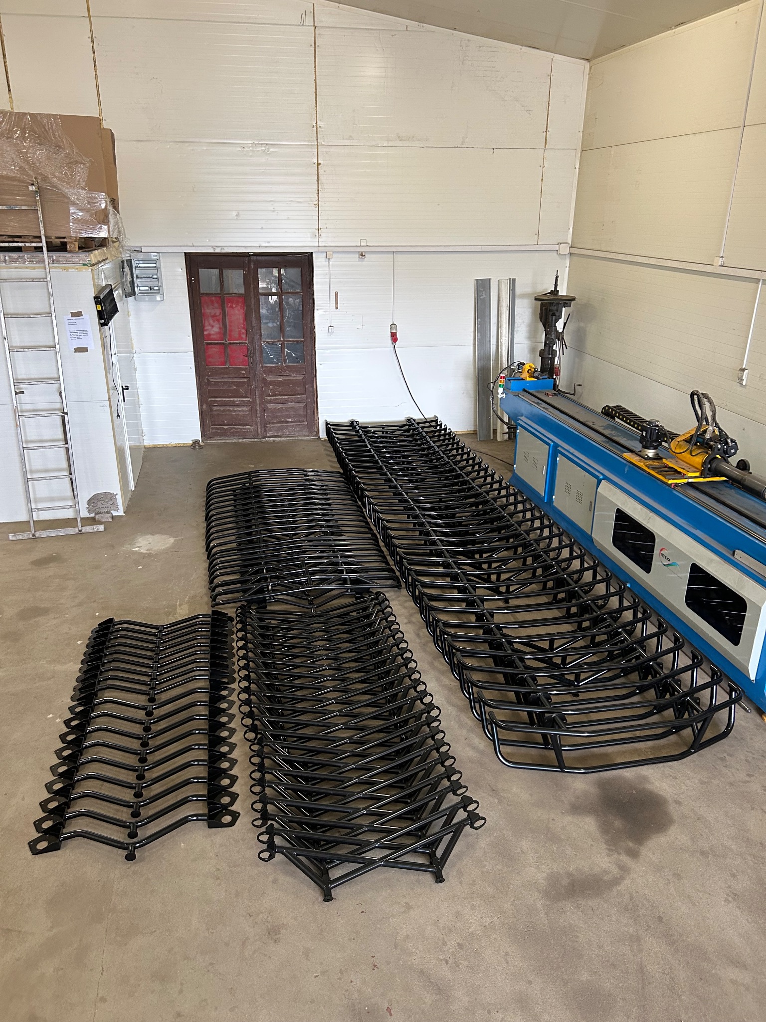 Partie czarnych, metalowych elementów konstrukcyjnych, prawdopodobnie do schodów, ułożone na podłodze hali obok maszyny do gięcia rur.