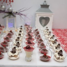 Crystal Catering & Events Kuriata Kacper - Rzędy deserów w szklanych naczyniach na białym stole z czerwonym bieżnikiem w śnieżynki, w tle lampion i dekoracje świąteczne.