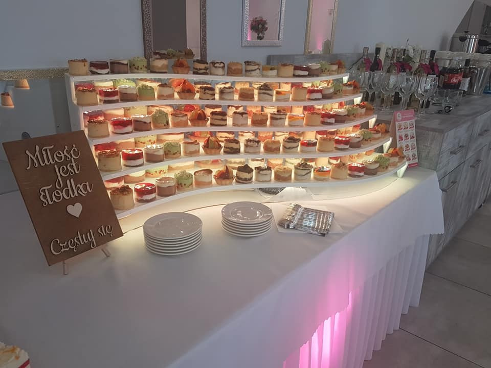 Stół z białym obrusem, na którym znajduje się wielopoziomowy regał z małymi deserami, obok stosy talerzy i drewniany napis 'Miłość jest słodka, Częstuj się'. W tle butelki wina i szklanki.