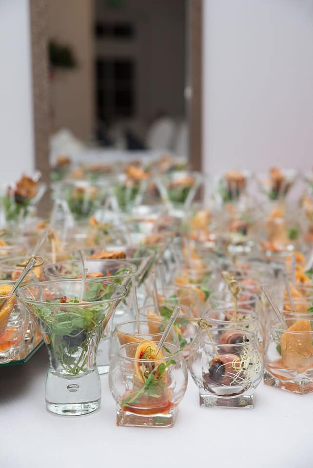 Szereg szklanych naczyń z apetycznymi przekąskami na białym stole, widoczne różne farsze i dodatki, w tle rozmyte wnętrze.