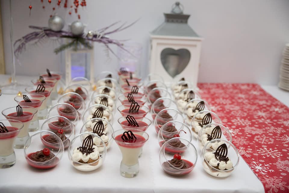 Rzędy deserów w szklanych naczyniach na białym stole z czerwonym bieżnikiem w śnieżynki, w tle lampion i dekoracje świąteczne.