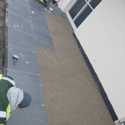 BRUK BUD PAWEŁ ŚLIWA - Montaż ciemnoszarych płyt kamiennych na tarasie przy białej ścianie budynku z oknem, widoczne fugi krzyżykowe i narzędzia pracy