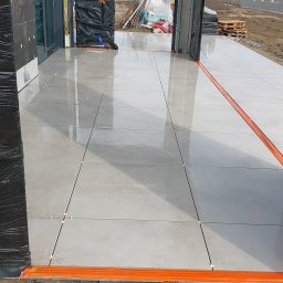 BRUK BUD PAWEŁ ŚLIWA - Jasnoszare płytki tarasowe ułożone na zewnątrz, widoczne pomarańczowe listwy zabezpieczające i krzyżyki dystansowe między płytkami, w tle ziemia i materiały budowlane.