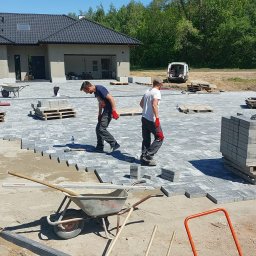 BRUK BUD PAWEŁ ŚLIWA - Dwóch mężczyzn w trakcie układania szarej kostki brukowej na dużej powierzchni przed domem z ciemnym dachem; widoczne palety z kostką, taczka i częściowo ułożona nawierzchnia.