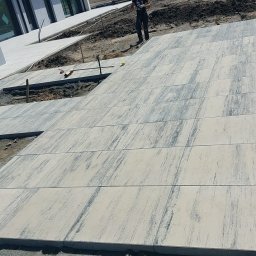 BRUK BUD PAWEŁ ŚLIWA - Świeżo ułożona, jasna kostka brukowa z szarymi smugami na posesji, widoczni pracownicy i narzędzia w tle, teren przygotowany pod dalsze prace.