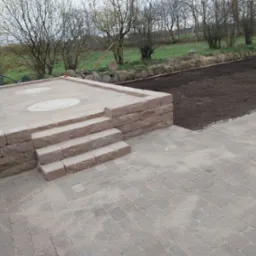 Ułożona kostka brukowa w odcieniach szarości i brązu, tworząca taras z niskim murkiem i schodami; w tle przygotowany teren pod trawnik.