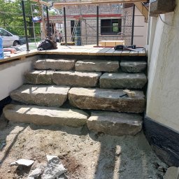 BRUK BUD PAWEŁ ŚLIWA - Kamienne schody prowadzące na drewniany taras w trakcie budowy, widoczne narzędzia i materiały budowlane, w tle rusztowanie i budynek w trakcie prac wykończeniowych.