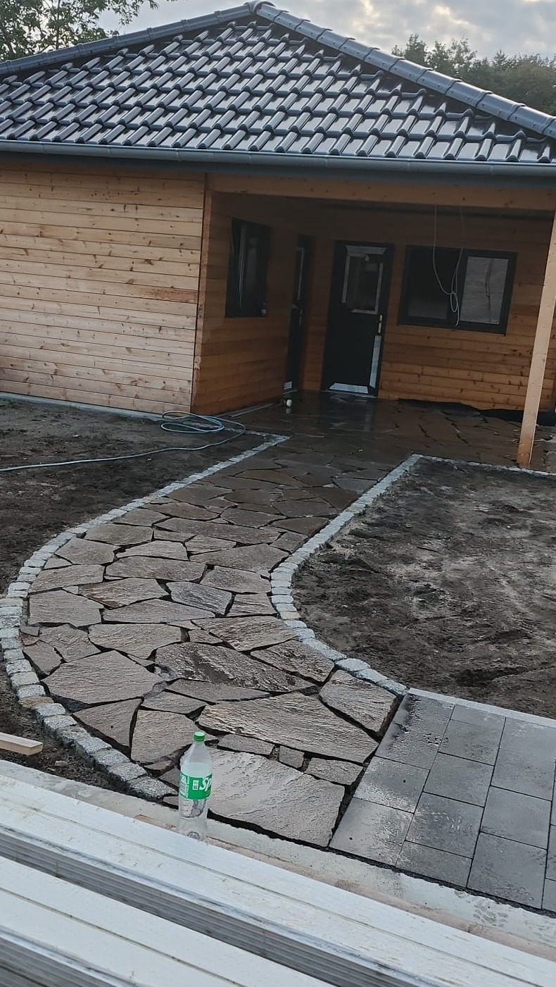 Ścieżka z nieregularnych kamieni prowadząca do drewnianego domku z ciemną stolarką, obrzeża z kostki granitowej, butelka wody przy krawędzi ścieżki, częściowo ułożona kostka brukowa w tle.