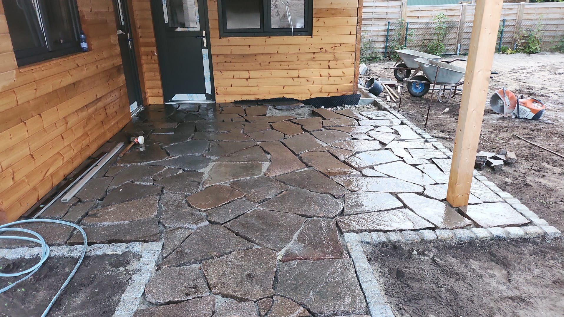 Nieregularny taras z kamienia naturalnego w trakcie budowy przy drewnianym domku, widoczne narzędzia i materiały budowlane, obrzeże z kostki granitowej.