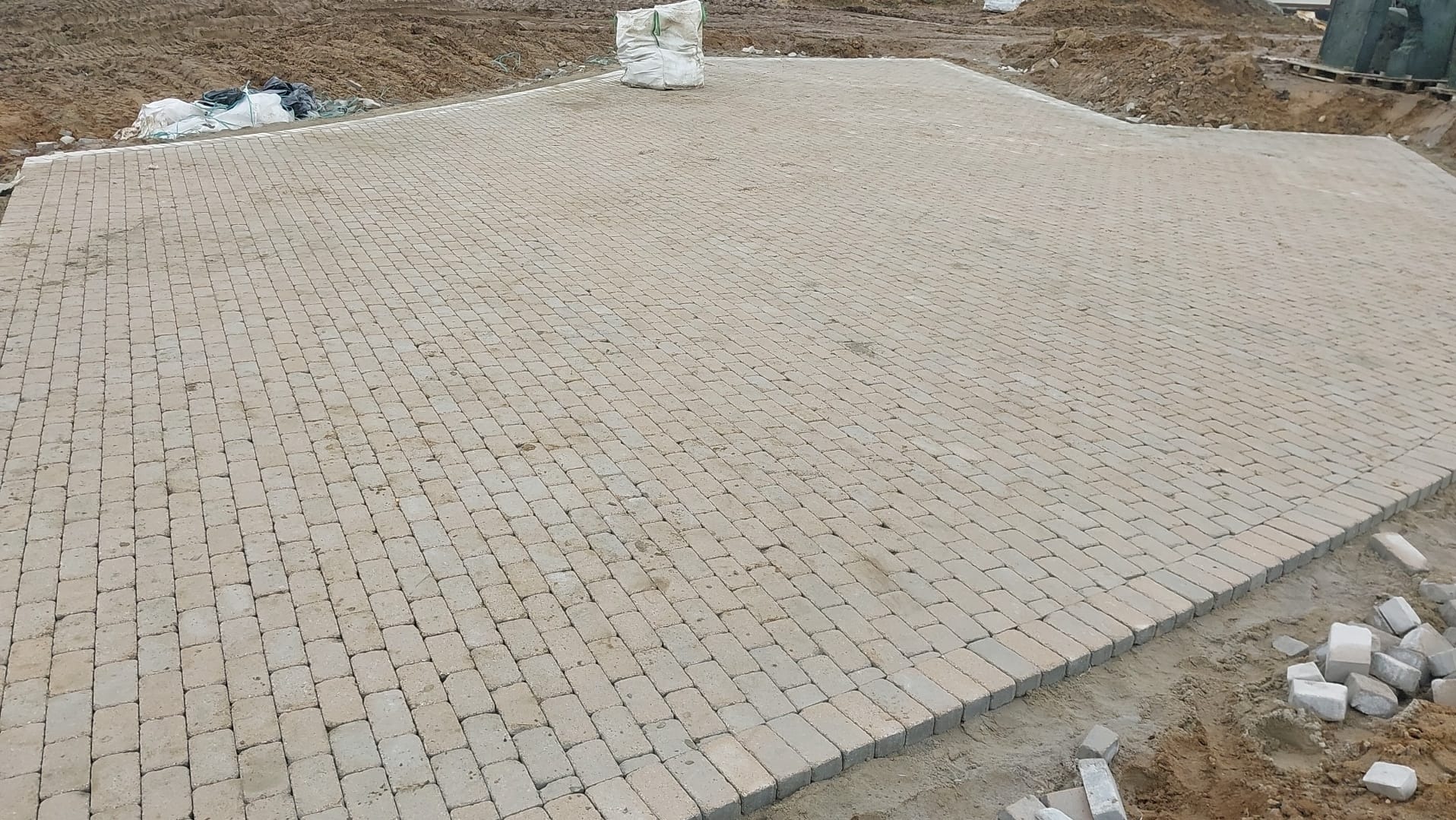 Niedokończona powierzchnia z beżowej kostki brukowej ułożonej w regularny wzór, z workami materiałów budowlanych i stertą kostek w tle.