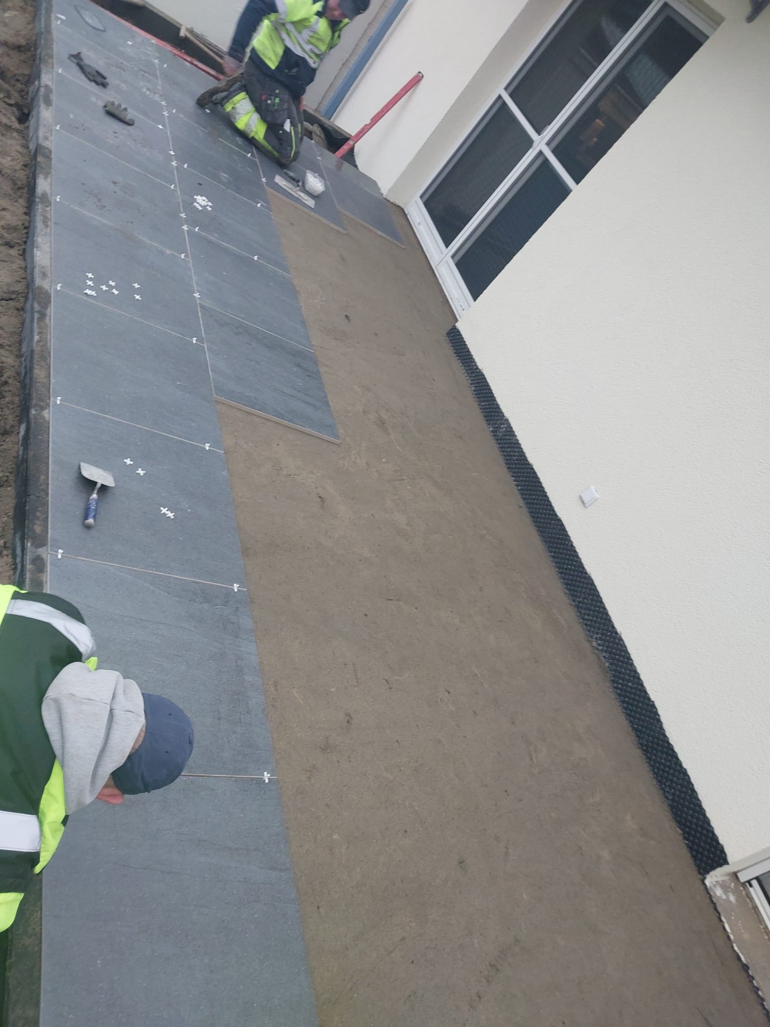 Montaż ciemnoszarych płyt kamiennych na tarasie przy białej ścianie budynku z oknem, widoczne fugi krzyżykowe i narzędzia pracy