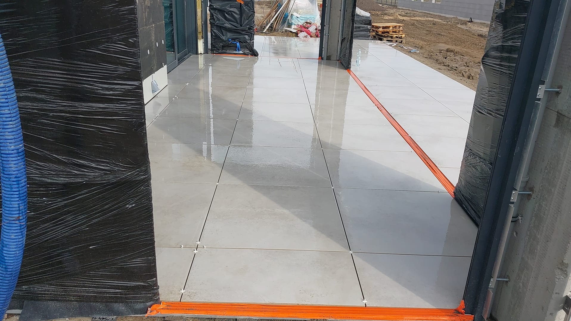 Jasnoszare płytki tarasowe ułożone na zewnątrz, widoczne pomarańczowe listwy zabezpieczające i krzyżyki dystansowe między płytkami, w tle ziemia i materiały budowlane.