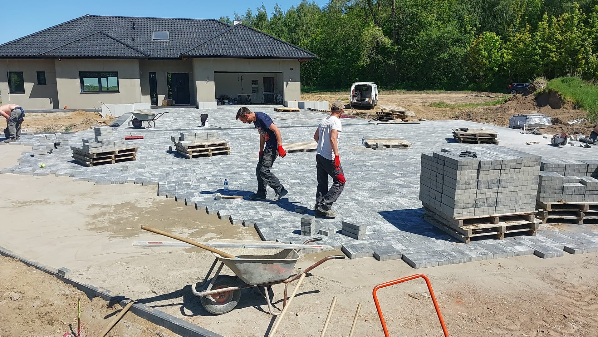 Dwóch mężczyzn w trakcie układania szarej kostki brukowej na dużej powierzchni przed domem z ciemnym dachem; widoczne palety z kostką, taczka i częściowo ułożona nawierzchnia.