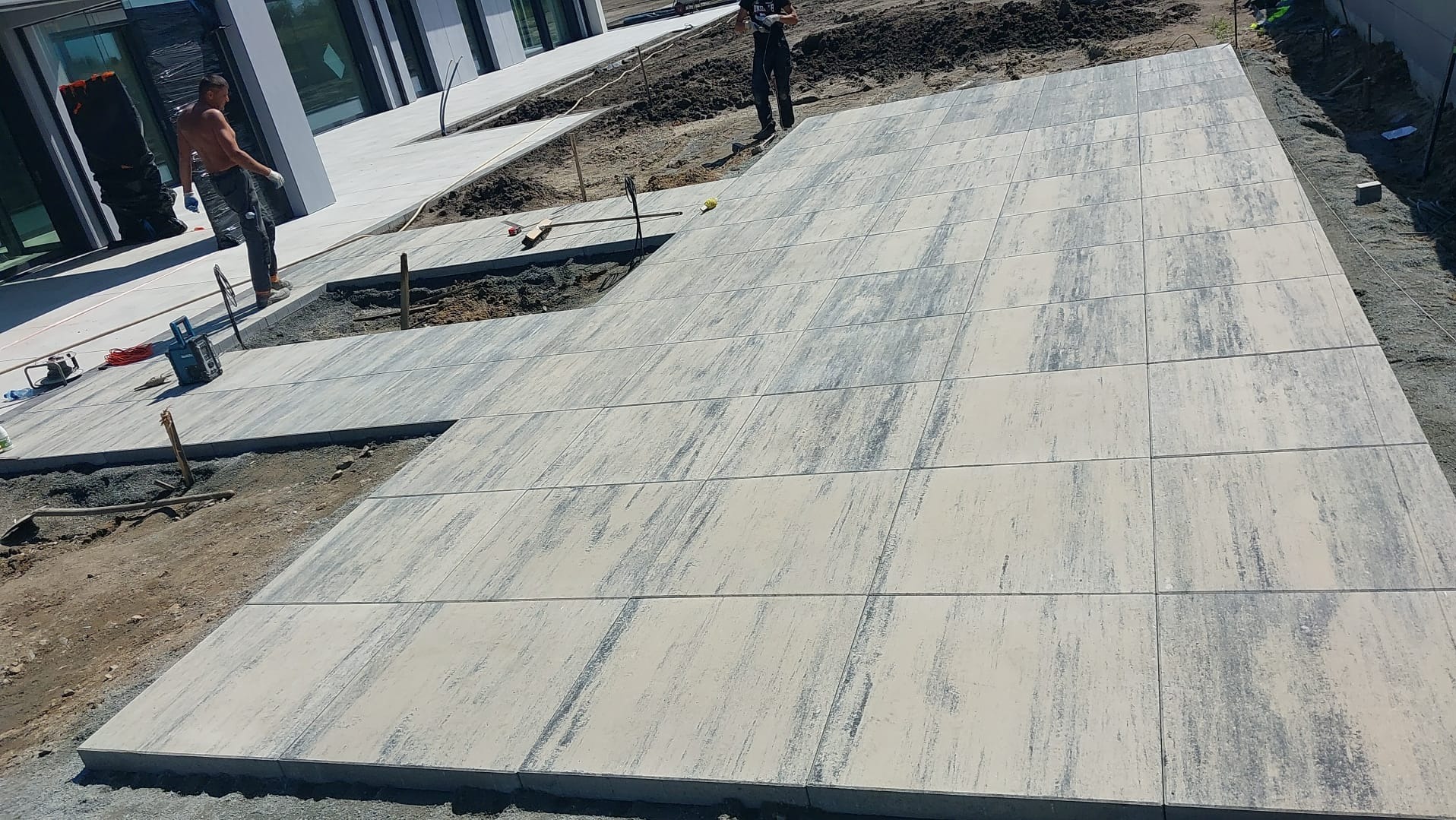 Świeżo ułożona, jasna kostka brukowa z szarymi smugami na posesji, widoczni pracownicy i narzędzia w tle, teren przygotowany pod dalsze prace.