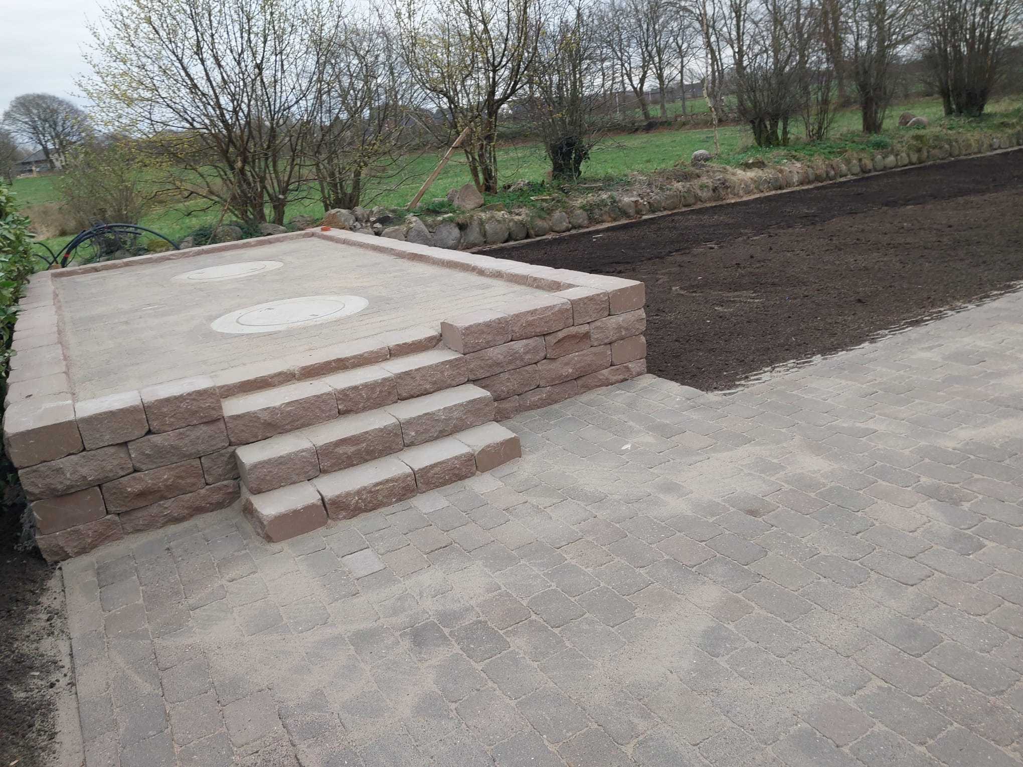 Ułożona kostka brukowa w odcieniach szarości i brązu, tworząca taras z niskim murkiem i schodami; w tle przygotowany teren pod trawnik.