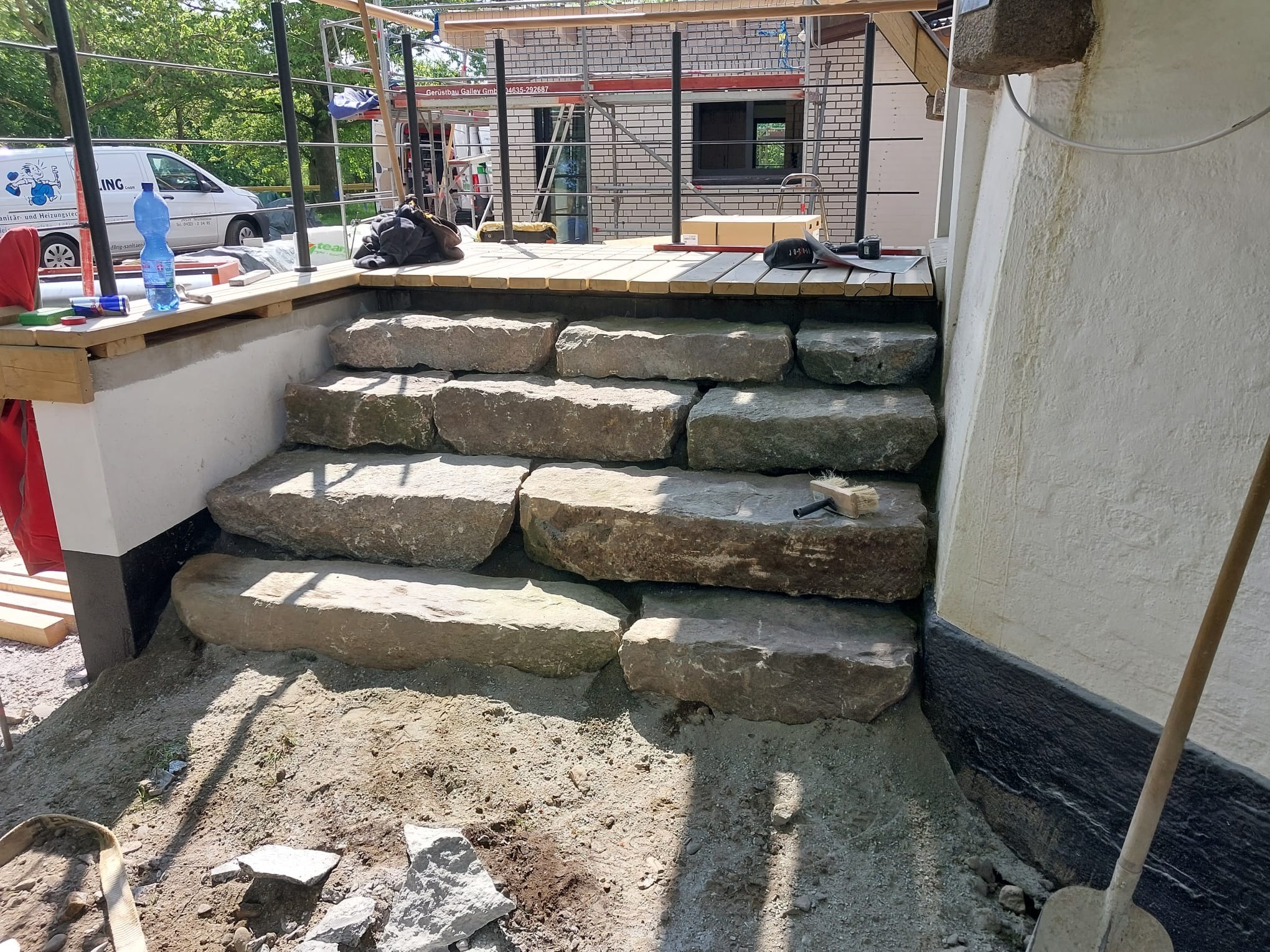 Kamienne schody prowadzące na drewniany taras w trakcie budowy, widoczne narzędzia i materiały budowlane, w tle rusztowanie i budynek w trakcie prac wykończeniowych.