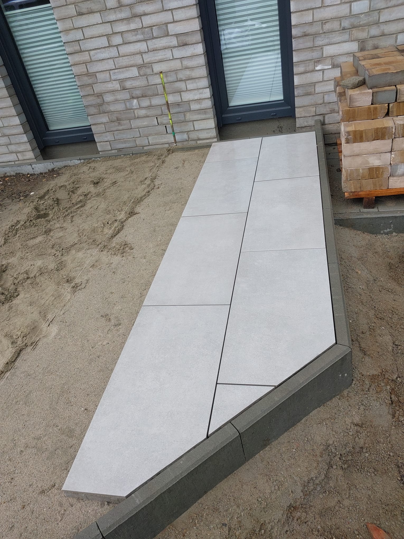 Układanie jasnoszarych płyt tarasowych na piaskowym podłożu, obramowanych ciemnoszarą kostką, widok na fragment elewacji z oknami.