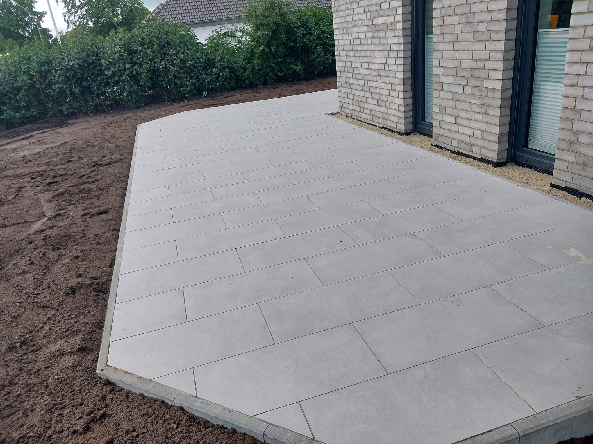 Nowoczesny taras z jasnoszarych płyt betonowych, obrzeża z szarej kostki, świeżo przygotowany teren pod trawnik, fragment elewacji z jasnej cegły.