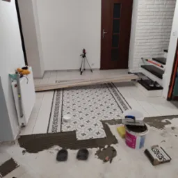 Układanie wzorzystych płytek podłogowych w przedpokoju, widoczny laser krzyżowy, wiadro z klejem Rapid, poziomica na grzejniku i fragment schodów z butami.