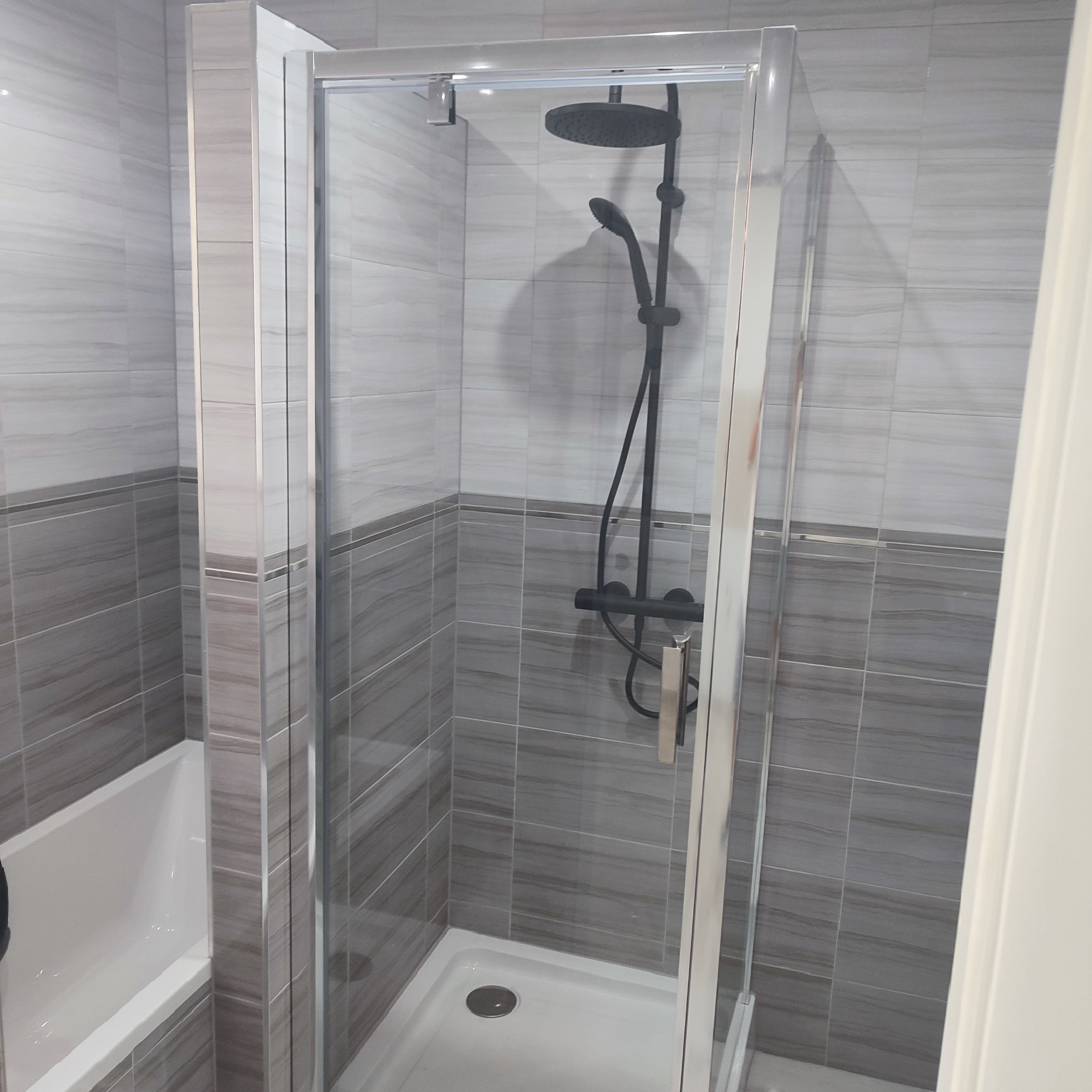 Nowoczesna kabina prysznicowa z czarną armaturą na tle płytek w odcieniach szarości, widok z boku z fragmentem wanny.