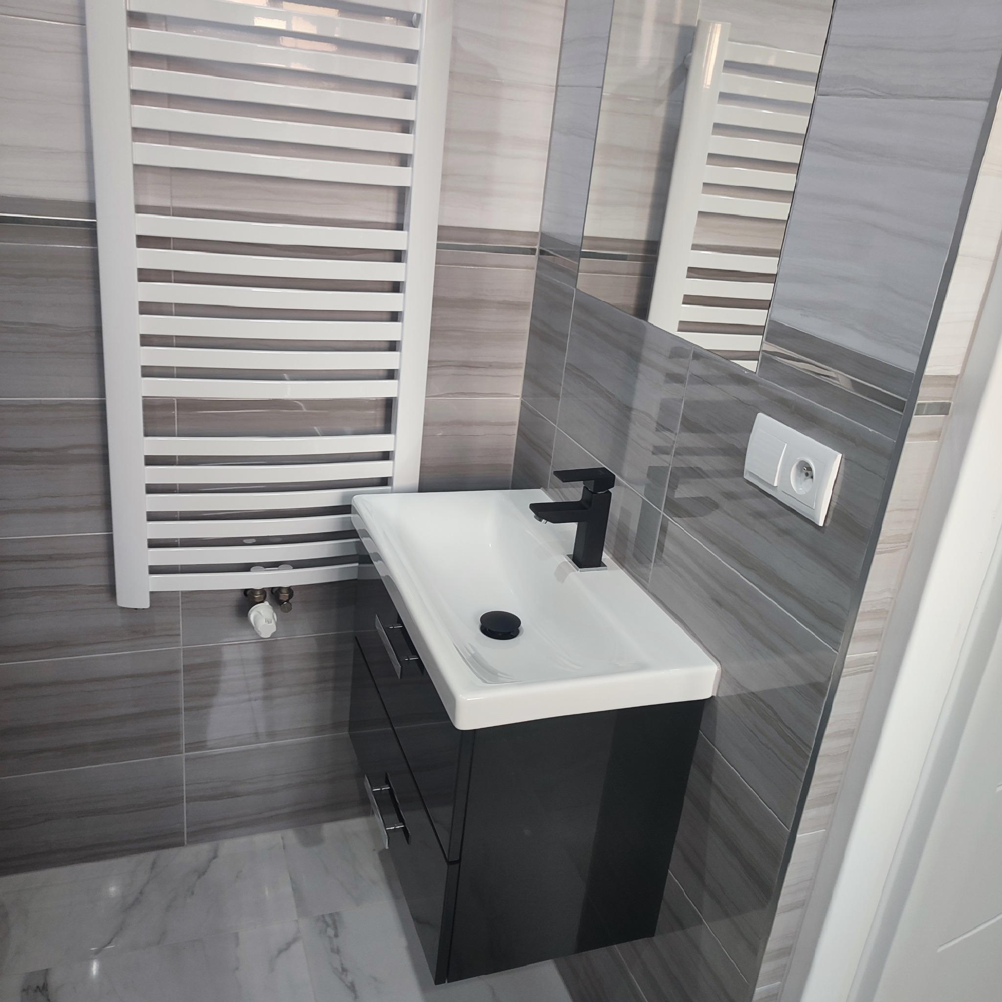 Nowoczesna łazienka w Wojniczu: szare płytki imitujące marmur, biała umywalka z czarną armaturą na szafce z szufladami, biały grzejnik drabinkowy i lustro.