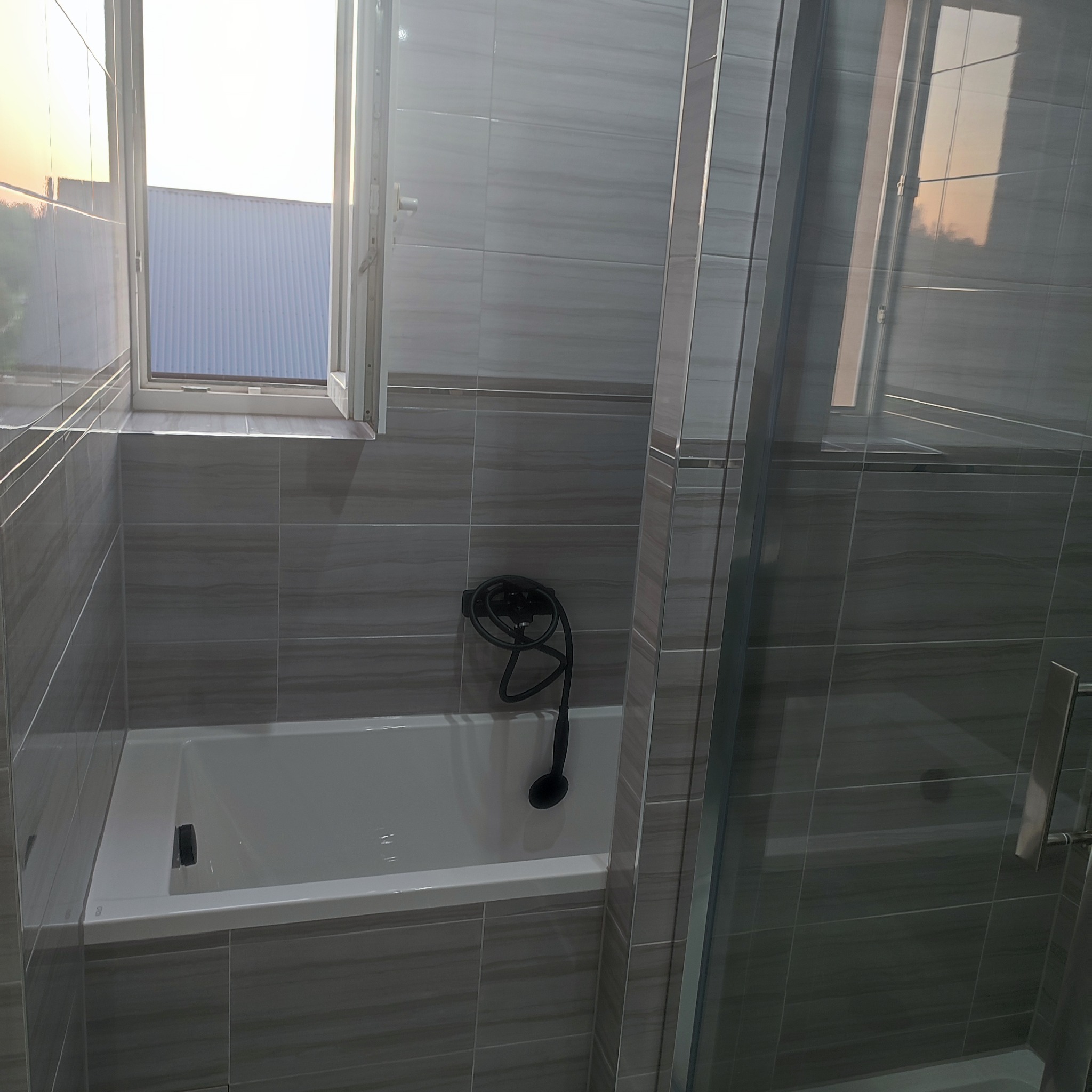 Nowoczesna łazienka z szarymi płytkami imitującymi drewno, białą wanną i czarną armaturą, widok na zachodzące słońce za oknem.