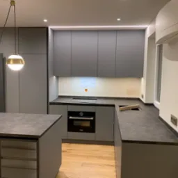 Szara, nowoczesna kuchnia z wyspą, oświetleniem LED podszafkowym i wiszącymi lampami kulami. Widoczne ślady po remoncie na ścianie nad blatem.