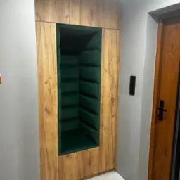 Wnęka w zabudowie z drewnopodobnej płyty, wyłożona ciemnozieloną, pikowaną tapicerką, tworząca miejsce do siedzenia lub czytania.