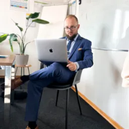 Mężczyzna w niebieskim garniturze i okularach siedzi na krześle z laptopem Apple, w tle biała tablica i roślina doniczkowa.