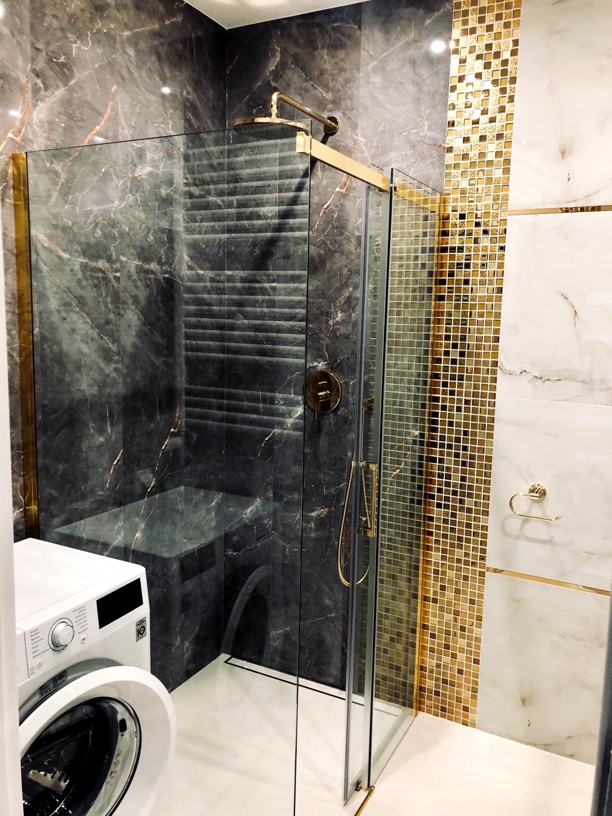 Nowoczesna łazienka z kabiną prysznicową typu walk-in wykończoną marmurem i złotymi akcentami, widoczna pralka z przodu po lewej stronie.