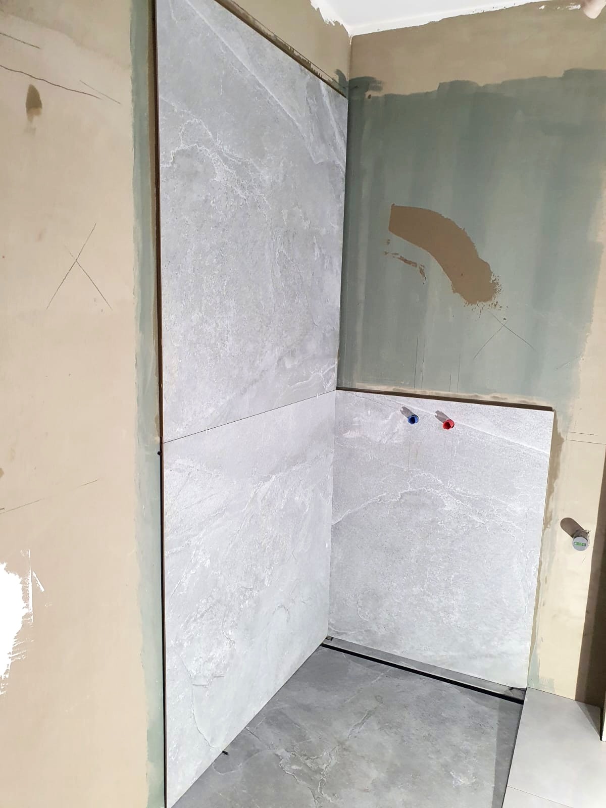 Montaż szarych płytek imitujących kamień w rogu łazienki, widoczne przyłącza wody z czerwonym i niebieskim oznaczeniem, ściany częściowo przygotowane pod malowanie.