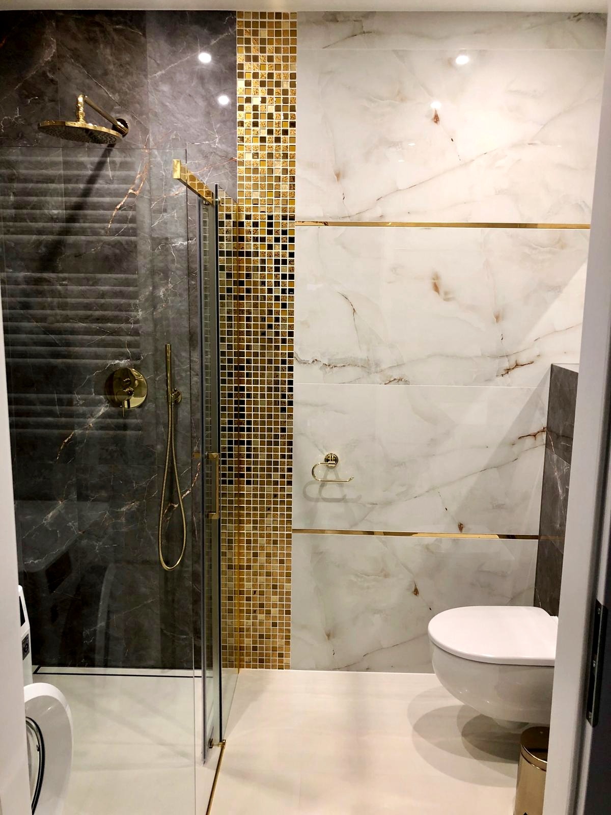 Elegancka łazienka z kabiną prysznicową typu walk-in, wykończona płytkami imitującymi marmur w odcieniach szarości i bieli, z akcentami złotych mozaik i armatury, widok na toaletę i fragment pralki.